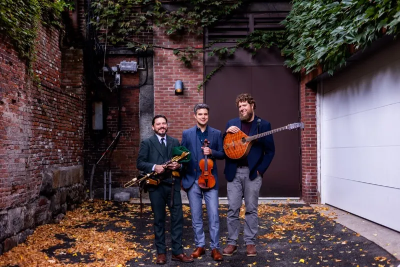 Joey Abarta, Nathan Gourley, and Owen Marshall standing in a brick alley with autumn leaves, holding their instruments
